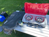 Essential gear for RV rental adventures: Coleman camping stove, propane, and a folding table on green grass.