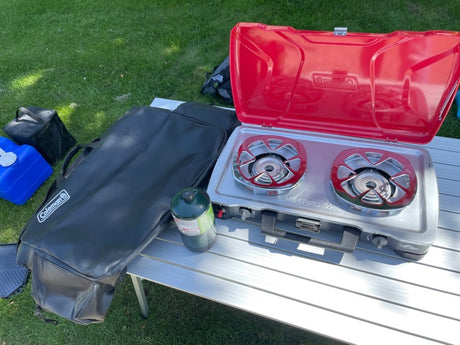 Essential gear for RV rental adventures: Coleman camping stove, propane, and a folding table on green grass.