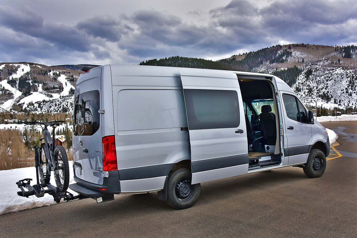 Silver RV rental van with bikes on hitch rack in a snowy mountain landscape. Side door open