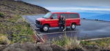 A couple with their red RV rental van in a rocky mountain parking lot, with distant islands under a cloudy sky.