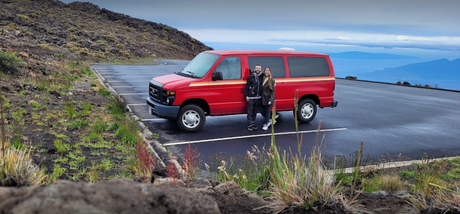 A couple with their red RV rental van in a rocky mountain parking lot, with distant islands under a cloudy sky.