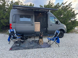 A gray camper van RV rental parked on gravel with an outdoor setup, including chairs, a table, and a long-haired German Shepherd dog.