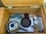 RV rental: Well-organized camp kitchen essentials in a built-in wooden storage unit, including stove, pans, mugs, and utensils.