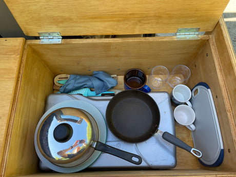 RV rental: Well-organized camp kitchen essentials in a built-in wooden storage unit, including stove, pans, mugs, and utensils.