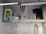 An RV rental kitchen with an open cabinet storing paper towels, a coffee maker, and dishes. A sink faucet and warning labels are below.