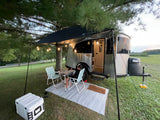 Enjoying an RV rental. A person dines outside a modern travel trailer under an awning with string lights at dusk.