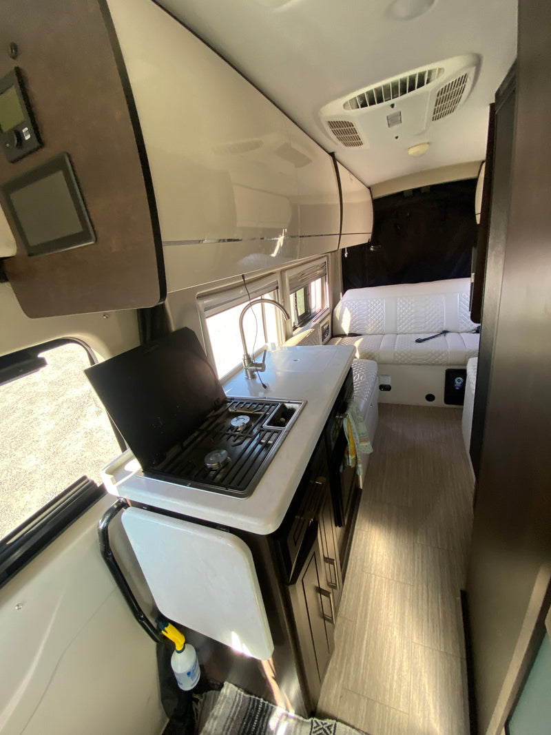 Modern RV rental interior: kitchen with stove, sink, and white quilted sofa; bright windows, overhead AC. — photo 3
