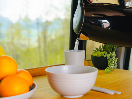Pouring water from a black kettle into a cup inside an RV rental. Fresh oranges and a plant sit on the wooden counter by a window.