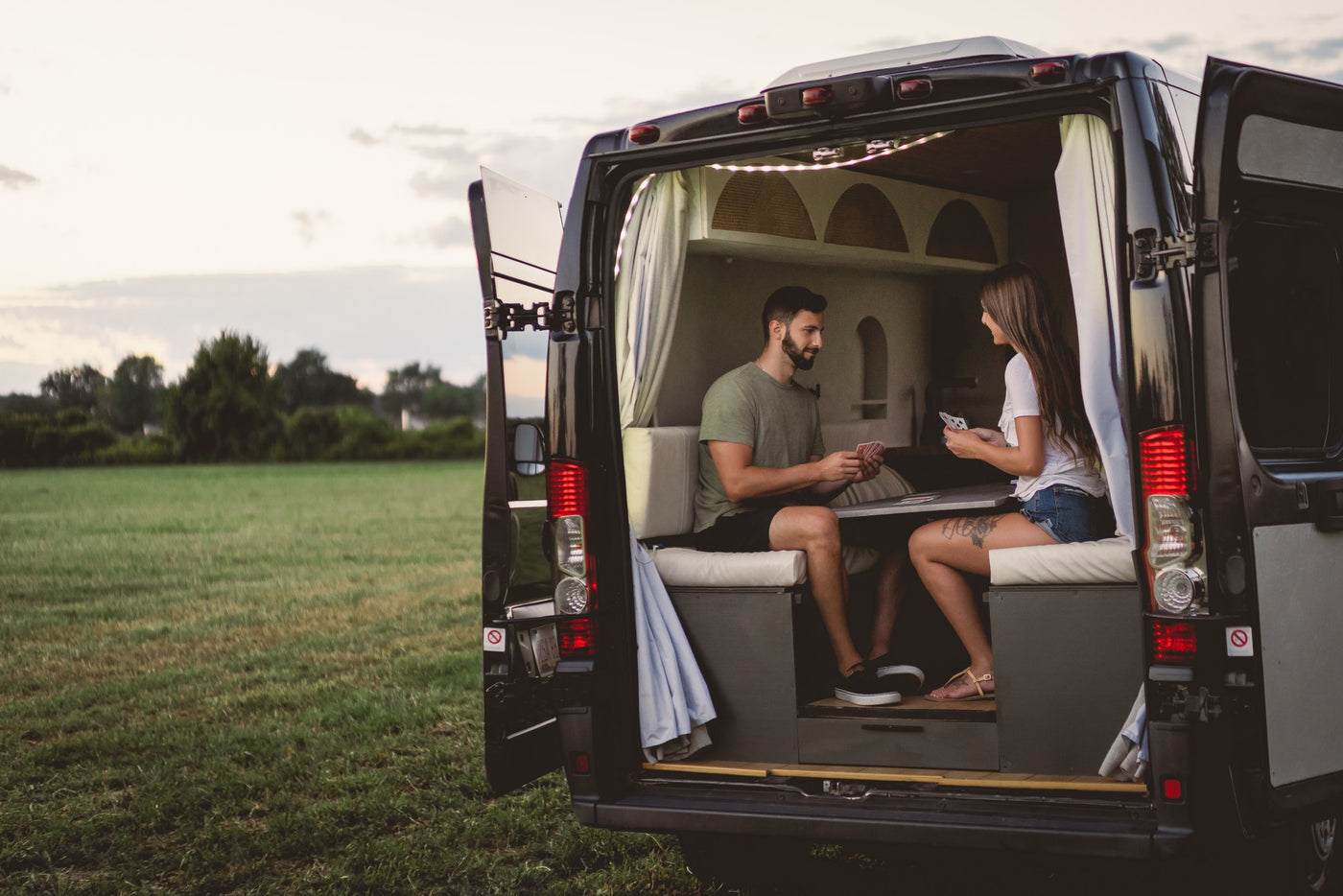 A couple plays cards inside their rv rental van, parked with its back doors open to a wide, grassy field. — photo 12