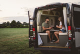 A couple plays cards inside their rv rental van, parked with its back doors open to a wide, grassy field.