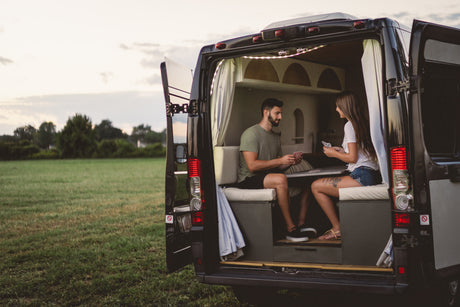 A couple plays cards inside their rv rental van, parked with its back doors open to a wide, grassy field.