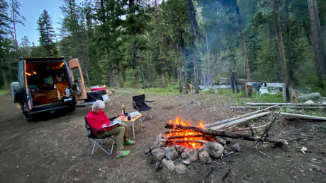 Serene RV rental camping: A person reads by a warm campfire in a lush forest with a river nearby, alongside their camper van.