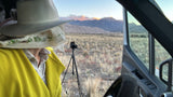 RV rental: Outdoors adventurer in hat enjoys scenic mountain views from inside, with camera on tripod for landscape photography.