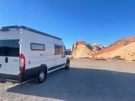 White RV rental van in a vast desert with stunning layered rock formations under a bright blue sky. Perfect for road trips.