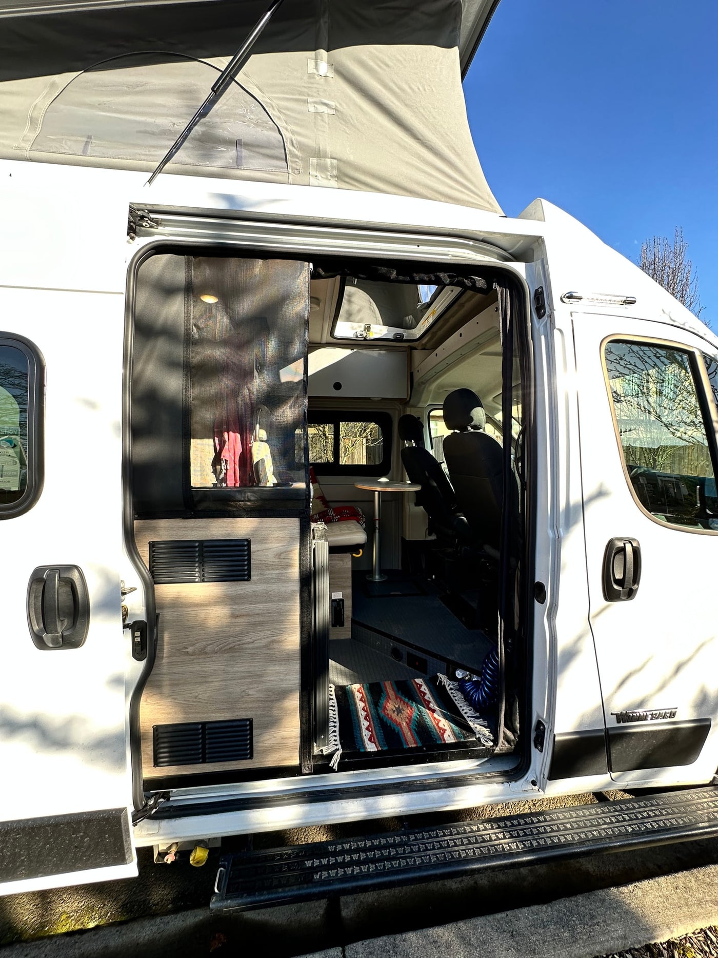 Open side door of an rv rental camper van reveals its pop-top roof and cozy interior with a table and rug. — photo 8