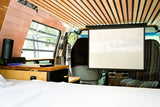 Spacious RV rental interior featuring a comfy bed, pull-down projector screen showing a basketball game, and custom wooden accents.