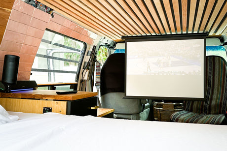 Spacious RV rental interior featuring a comfy bed, pull-down projector screen showing a basketball game, and custom wooden accents.