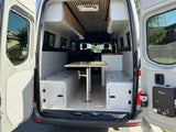 RV rental: Camper van interior reveals a versatile table, long storage benches, overhead cabinets, and rear water heater unit.