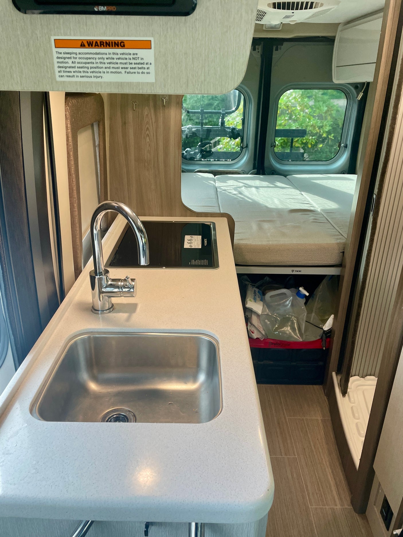 Inside a compact RV rental: full kitchen with sink & Dometic stovetop, rear bed, and storage. Safety warning on wall.