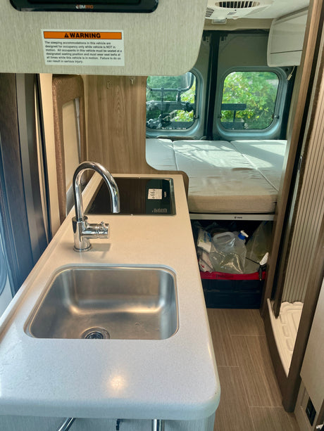 Inside a compact RV rental: full kitchen with sink & Dometic stovetop, rear bed, and storage. Safety warning on wall.