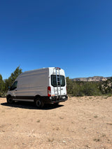 This white Ford Transit **RV rental** is ready for adventure, parked on a dirt road with majestic desert mountains.