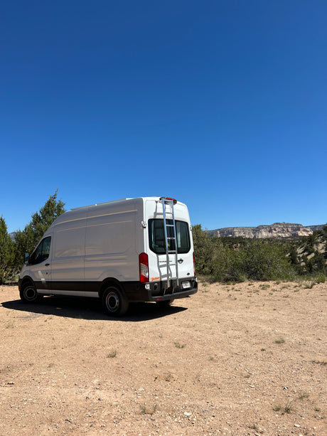 This white Ford Transit **RV rental** is ready for adventure, parked on a dirt road with majestic desert mountains.