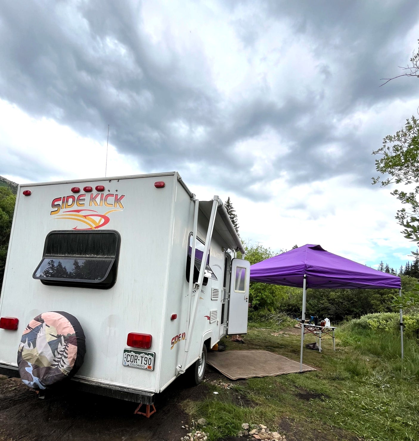 White "Side Kick" rv rental camper at a muddy campsite with a purple canopy, surrounded by green foliage under cloudy skies.