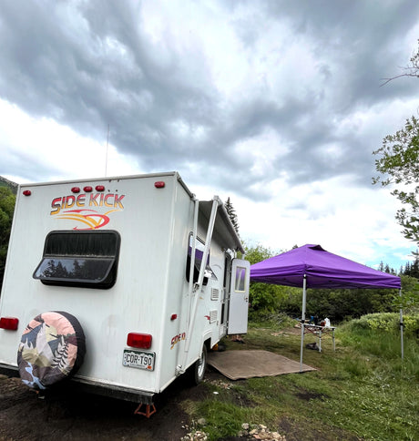 White "Side Kick" rv rental camper at a muddy campsite with a purple canopy, surrounded by green foliage under cloudy skies.