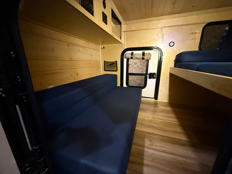 Cozy RV rental interior with warm wood paneling, featuring a blue bench bed, overhead storage, and a separate sleeping bunk.