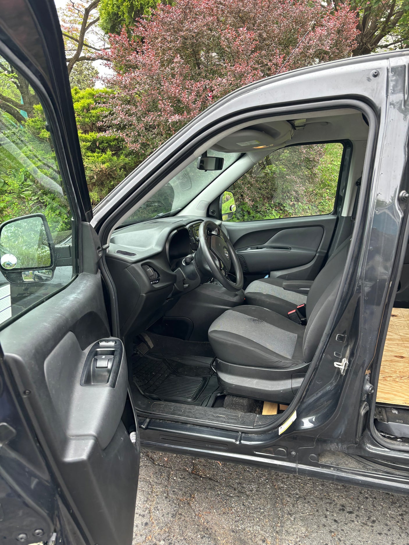 Inside a dark RV rental van: driver's seat, steering wheel, and open door showing lush greenery outside.
