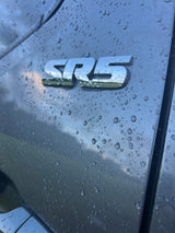 Close-up: Rain-covered grey vehicle with a shiny 'SRS' emblem reflecting cloudy skies. Ideal for an RV rental ad.