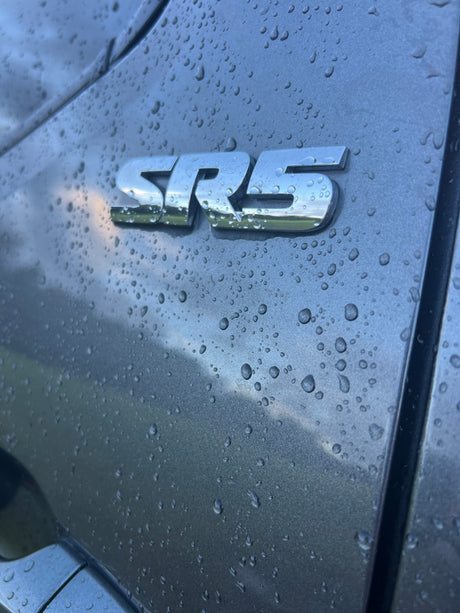 Close-up: Rain-covered grey vehicle with a shiny 'SRS' emblem reflecting cloudy skies. Ideal for an RV rental ad.