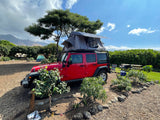 Experience outdoor adventure with this rv rental: a red Jeep with a deployed rooftop tent at a picturesque campsite. Mountains in background.