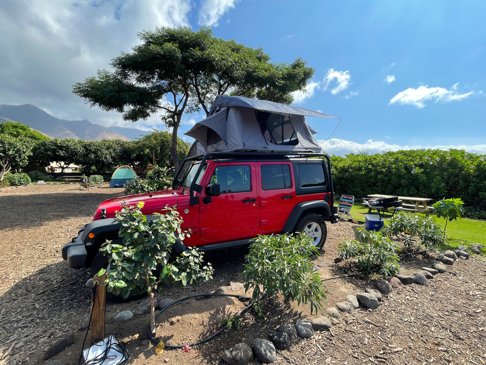 Experience outdoor adventure with this rv rental: a red Jeep with a deployed rooftop tent at a picturesque campsite. Mountains in background.