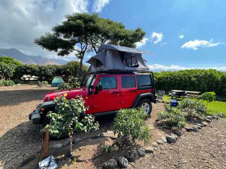Experience outdoor adventure with this rv rental: a red Jeep with a deployed rooftop tent at a picturesque campsite. Mountains in background.