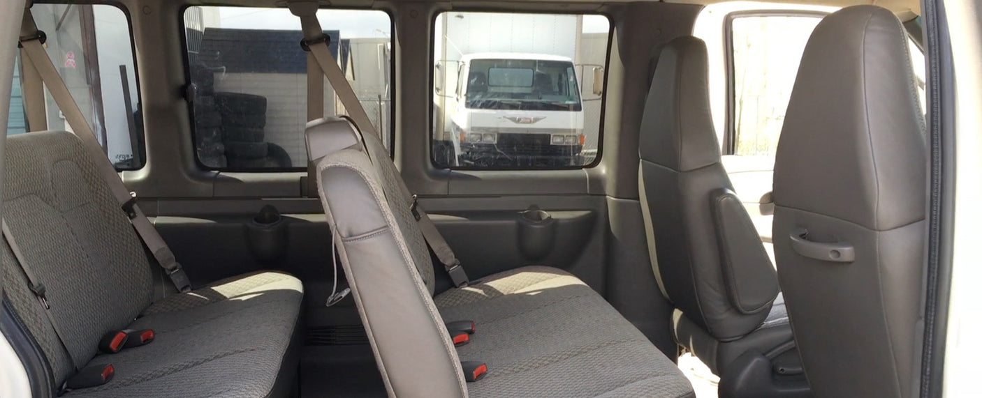 Inside a spacious RV rental van, showing rows of grey passenger seats. Outside, a white truck and industrial area are visible.