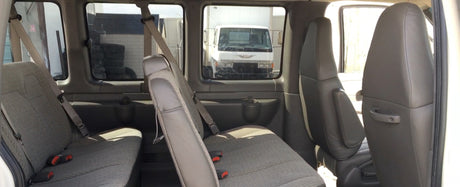 Inside a spacious RV rental van, showing rows of grey passenger seats. Outside, a white truck and industrial area are visible.