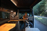 Man smiles, working from his RV rental mobile office, parked roadside by a forest. Ideal for digital nomads.