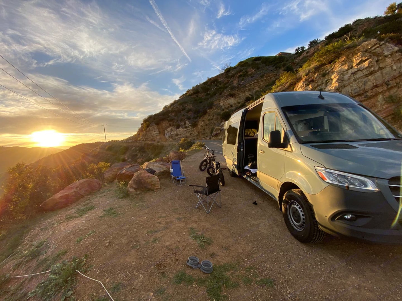 Scenic rv rental camper van parked on a mountain roadside at sunset, with chairs & a bike set up for a peaceful evening.