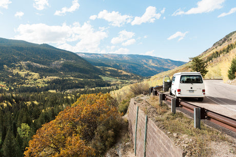 RV rental campervan on mountain road, overlooking an autumn valley with colorful trees and distant peaks.