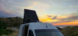 An RV rental with a deployed rooftop tent captures a vibrant sunset over hills and a distant lake. Scenic road trip!