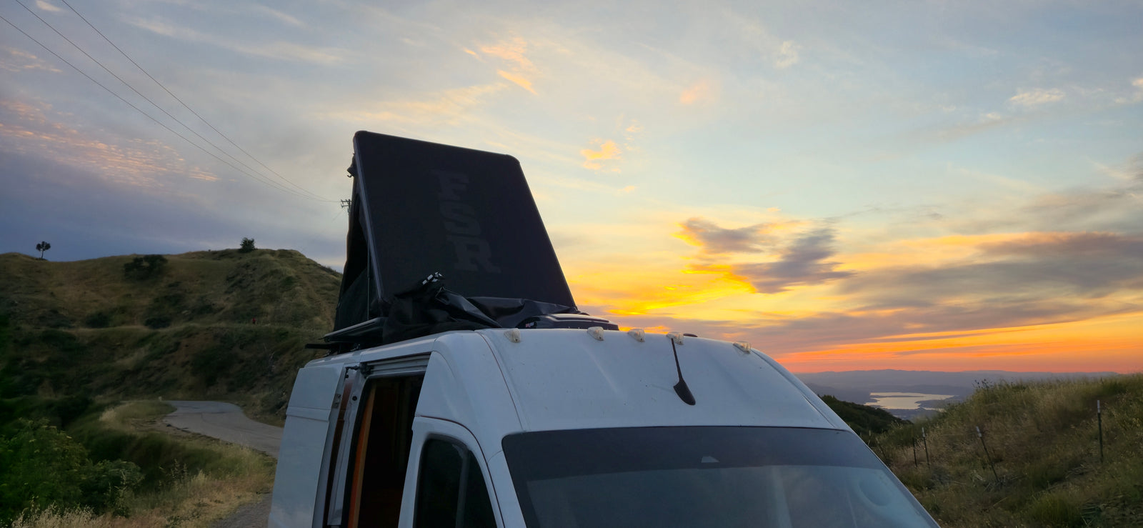 An RV rental with a deployed rooftop tent captures a vibrant sunset over hills and a distant lake. Scenic road trip!