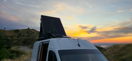 An RV rental with a deployed rooftop tent captures a vibrant sunset over hills and a distant lake. Scenic road trip!