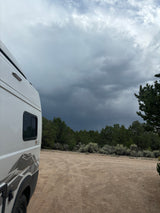 RV rental (Winnebago Solis) on a dirt road amidst trees, under a dramatic, stormy sky. Perfect for remote outdoor adventures.