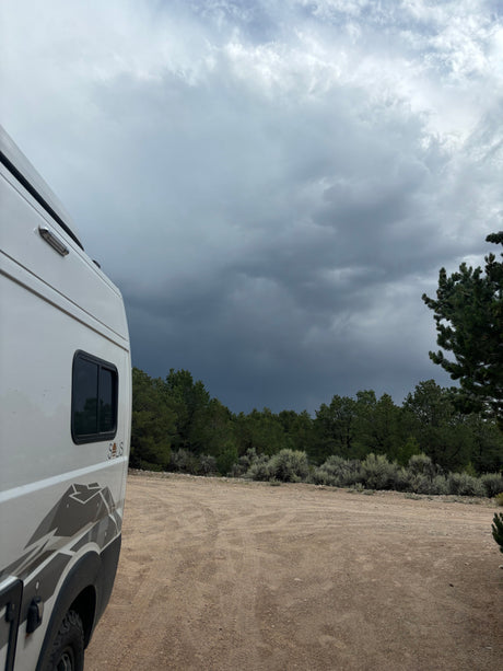 RV rental (Winnebago Solis) on a dirt road amidst trees, under a dramatic, stormy sky. Perfect for remote outdoor adventures.