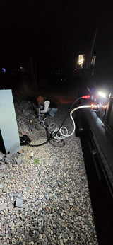 RV rental hookup at night. A person connects utilities to the camper on a gravel pad, illuminated by vehicle lights.