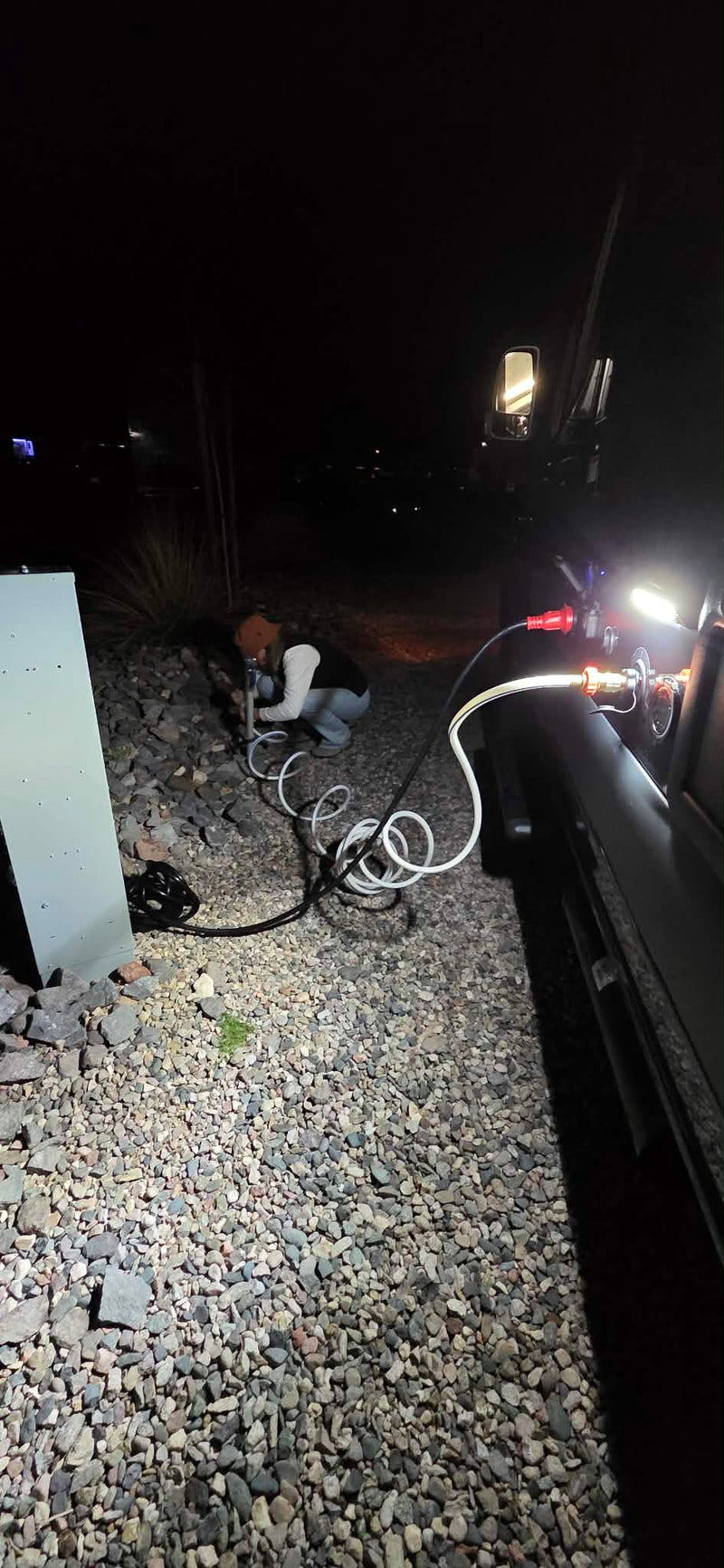 RV rental hookup at night. A person connects utilities to the camper on a gravel pad, illuminated by vehicle lights. — photo 17