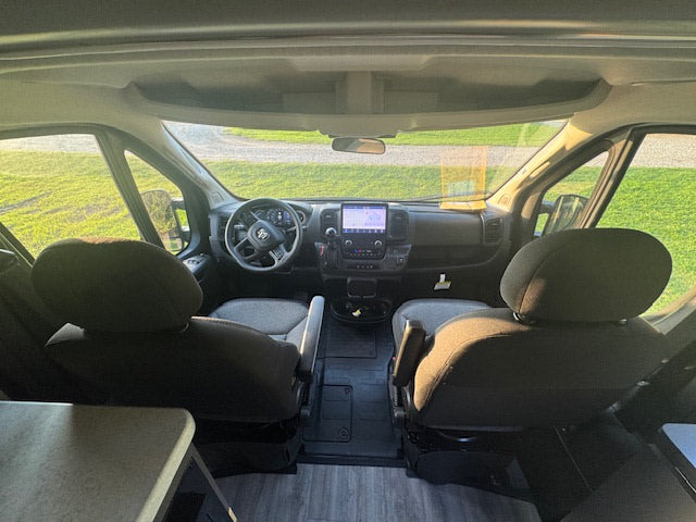 Inside an **rv rental**, looking forward past front seats to the dashboard, steering wheel, & windshield showing an outdoor field.