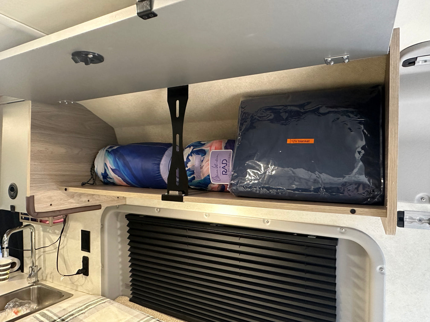 RV rental overhead storage with blankets and a 12V blanket, above a sink and window with blinds.