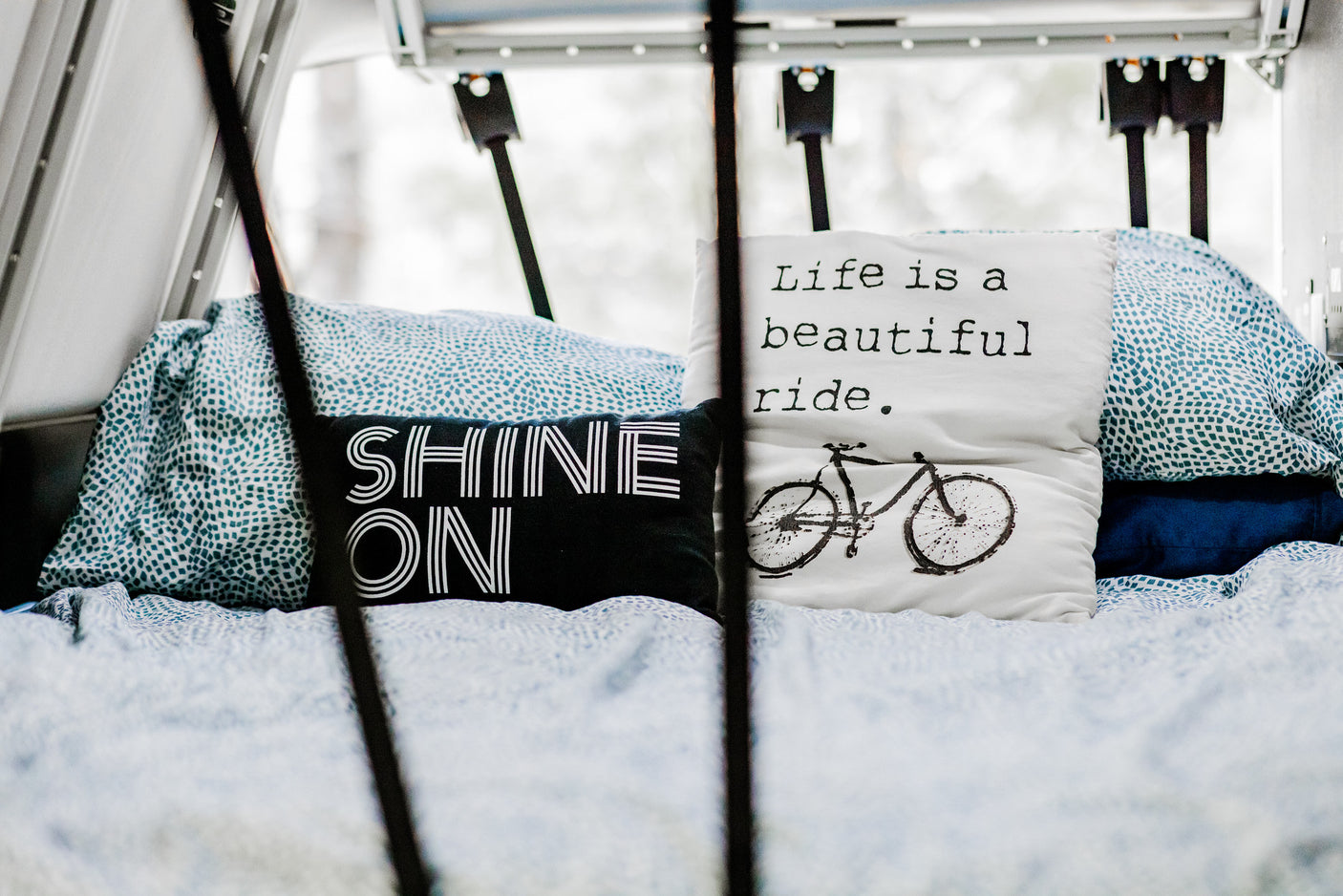 Inside an RV rental, a comfy bed with patterned linens holds pillows reading 'SHINE ON' and 'Life is a beautiful ride.'